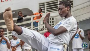 ciemnoskóra krótkowłosa kobieta wykonująca kopniecie podczas gry capoeira w roda a wokół dzieci oglądające wydarzenie ze schodów i balkonu a dookoła dzieci trenujące capoeira wszyscy kobieta ma na abada logo unicef